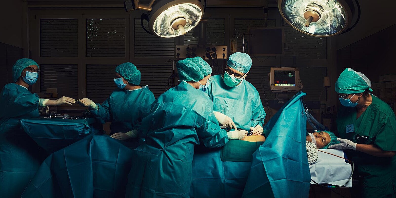 Unser Anspruch: Sie rundum hochwertig und zugleich menschlich zu versorgen. Ein Patient liegt auf dem OP-Tisch in einem modernen Operationssaal. In der Bildmitte operiert ein Arzt. Dabei spricht er mit einer Pflegekraft, die ihm assistiert. Rechts hält eine Anästhesistin den Kopf des Patienten fest. Im Hintergrund reicht eine weitere Pflegekraft einer Kollegin chirurgisches Instrument, dabei blicken sie sich an. Alle Personen tragen Mundschutz und türkisfarbene Schutzkleidung.