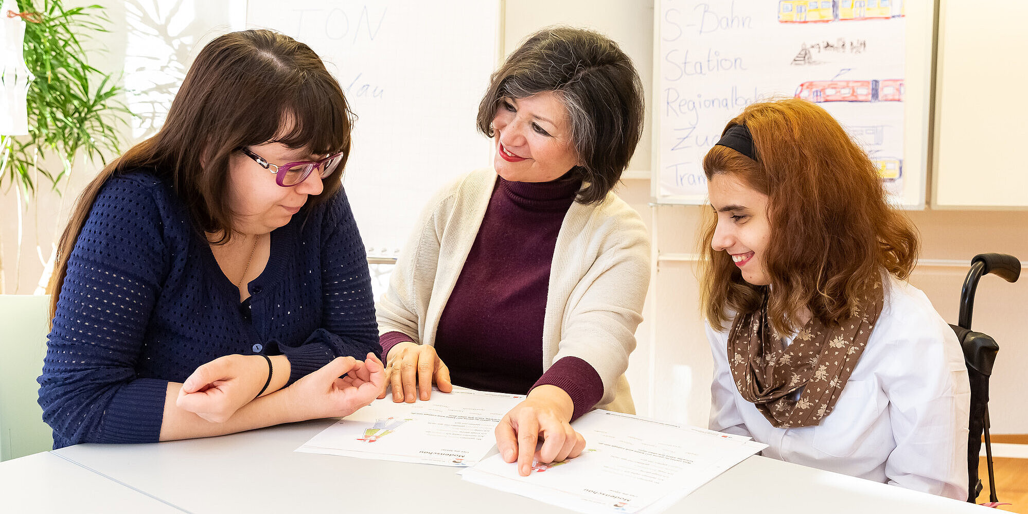 DiaLOG-IN – Sprache, Beratung, Coaching und Begleitung Drei Frauen arbeiten mit Arbeitsblättern an einem Tisch.